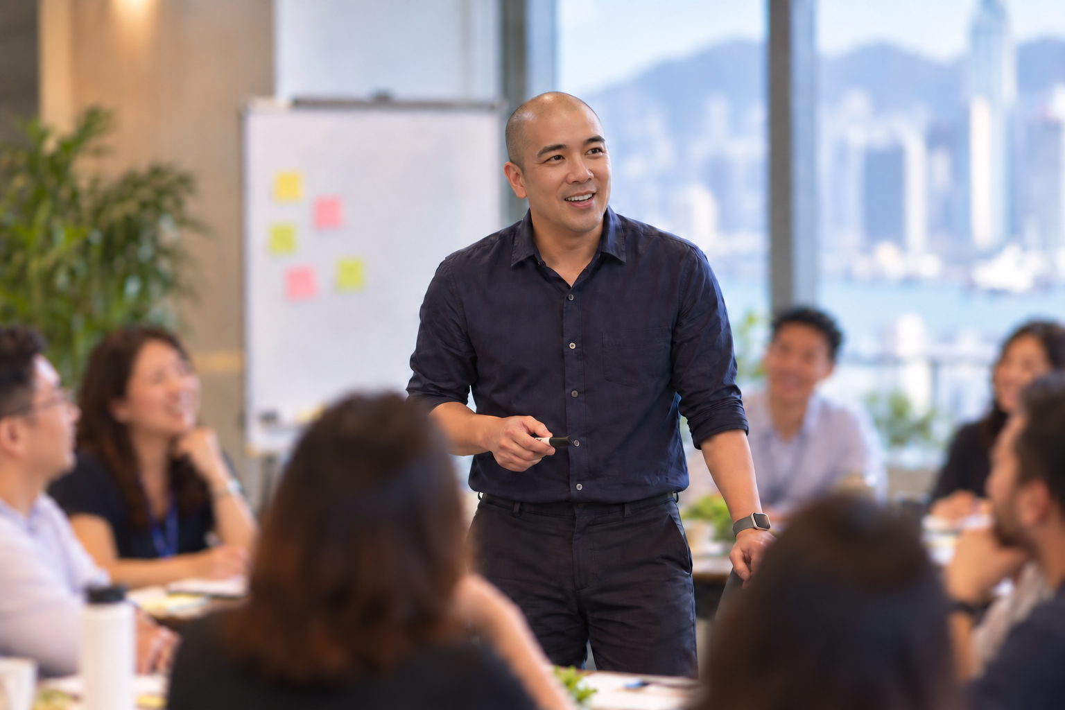 Norbert Woo leading a workshop with a group around a table, Hong Kong harbour beyond