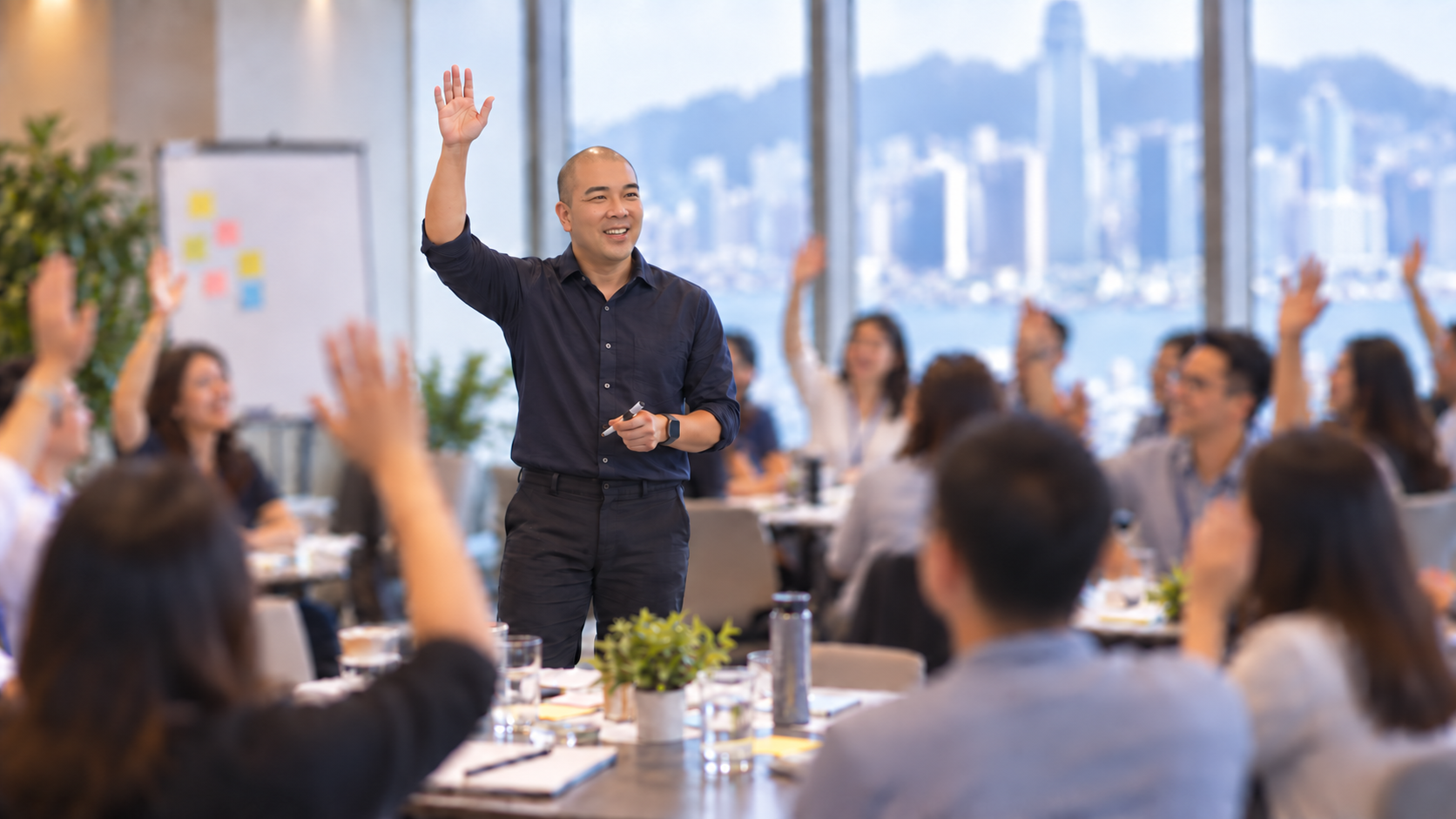 Norbert Woo leading a workshop with his hand raised, participants responding, Hong Kong harbour behind
